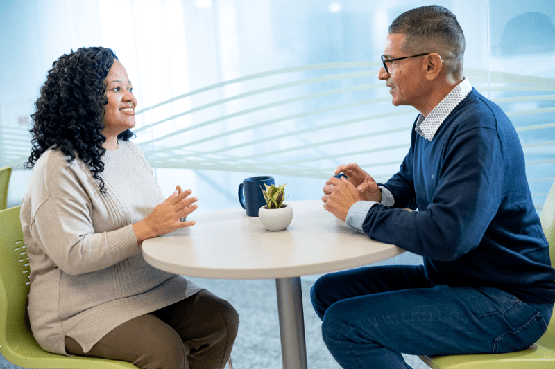 Two employees at a table with cups engaging in conversation