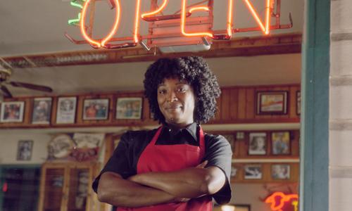Dueña de un restaurante parado frente a un letrero neÃ³n de ABIERTO en una ventana
